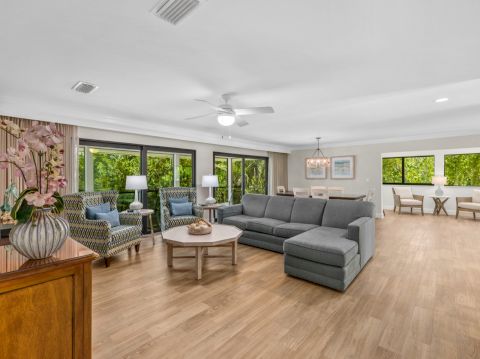Living room with a gray sectional sofa, two armchairs, a round coffee table, and large windows showing greenery
