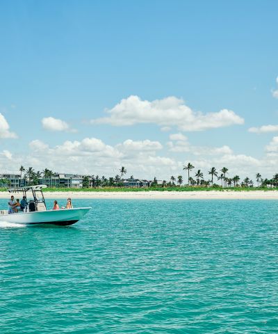 A speedboat cruises across turquoise water toward a sandy beach lined with palm trees and resort buildings under a blue sky.