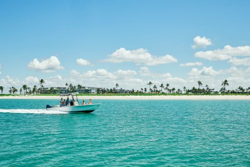 A speedboat cruises across turquoise water toward a sandy beach lined with palm trees and resort buildings under a blue sky.