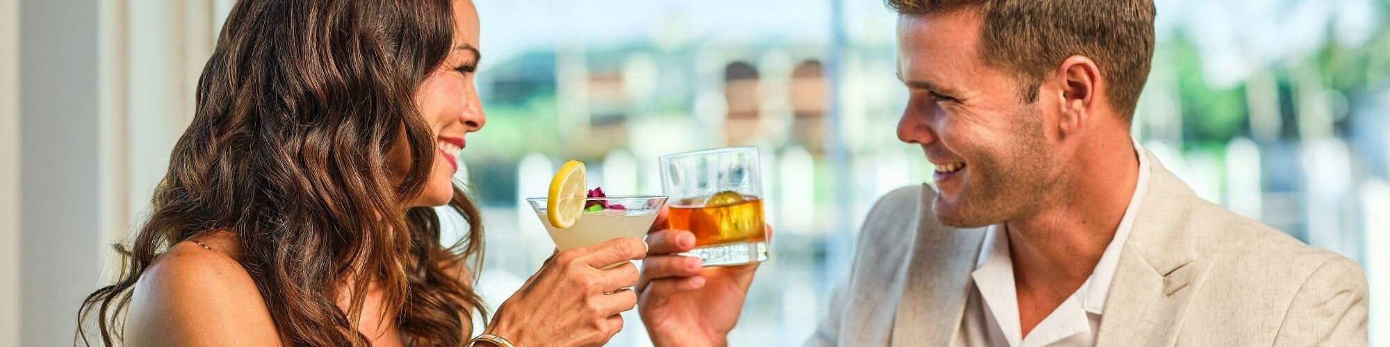 A couple clinks cocktail glasses with lemon and amber drink, smiling at each other in a bright bar.