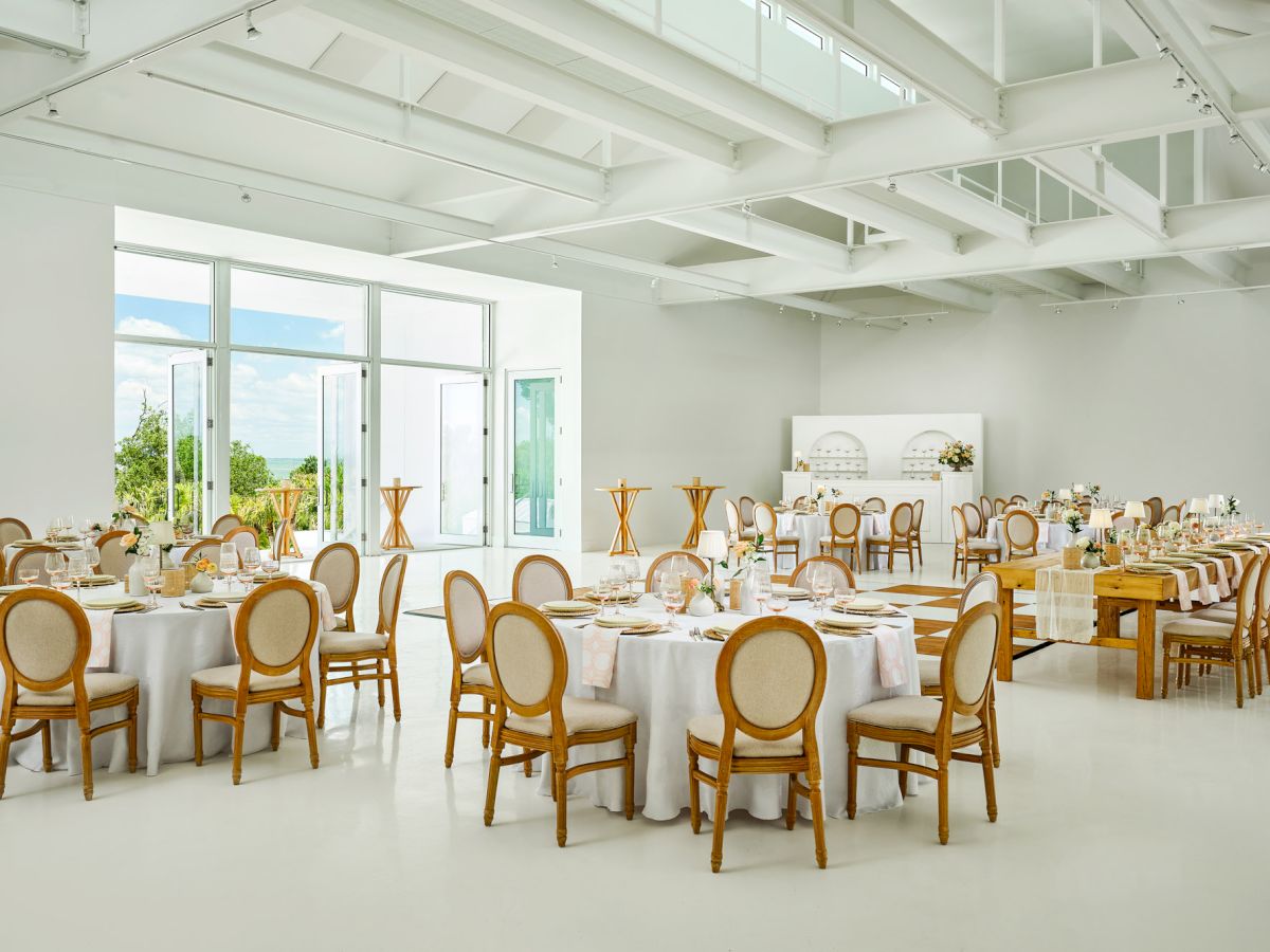A bright modern event hall set for a formal dinner, with long tables, place settings, flowers, and chairs by large windows.