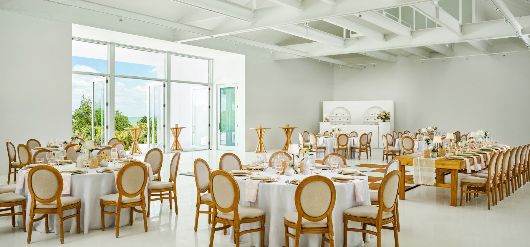 A bright modern event hall set for a formal dinner, with long tables, place settings, flowers, and chairs by large windows.