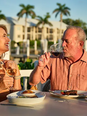 A couple on a sunny patio clinks and sips wine while dining, with a resort backdrop and palm trees behind them.