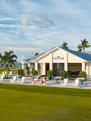 Sunny beachfront resort clubhouse labeled “THE CLUTCH,” with manicured lawn, palm trees, outdoor tables, and people relaxing under a blue sky.