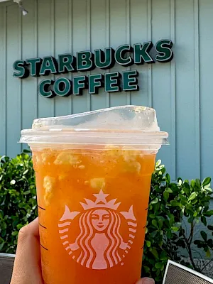 A hand holds a Starbucks iced orange drink outside a building with a “Starbucks Coffee” sign on the wall.