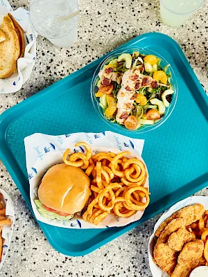 A cafeteria-style meal tray with salad, grilled chicken, a burger sandwich, fries, onion rings, cookies, and drinks.