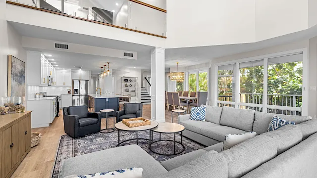 A modern open-plan living room with gray sofa, coffee table, and a bright kitchen with cabinets and stainless appliances.