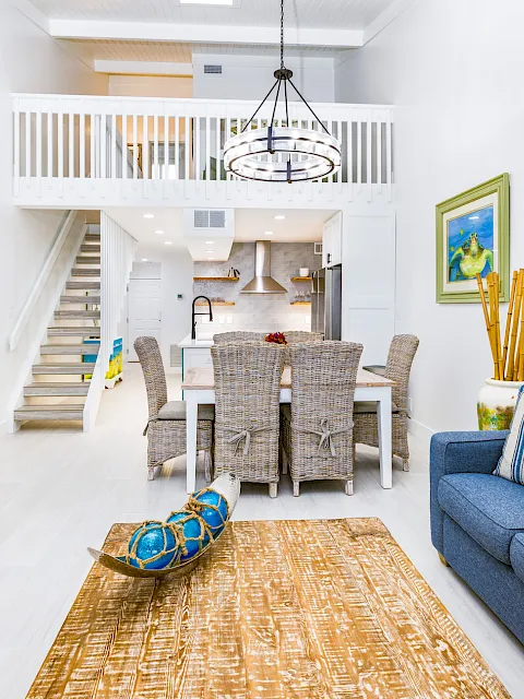 Bright coastal interior with hanging chandelier, wicker dining set, blue sofa, rug, staircase, and an open kitchen.