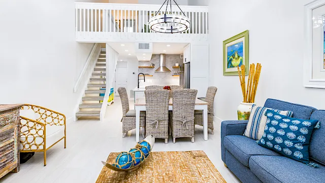 Bright coastal interior with hanging chandelier, wicker dining set, blue sofa, rug, staircase, and an open kitchen.