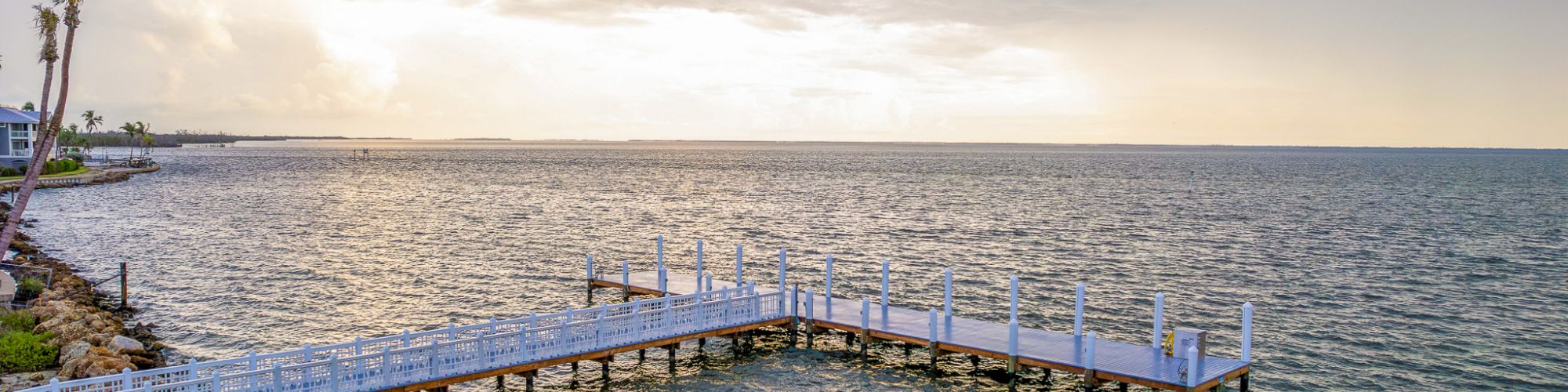 A white wooden dock with posts extends into calm ocean water under a cloudy sunset sky, with palm trees on the left.