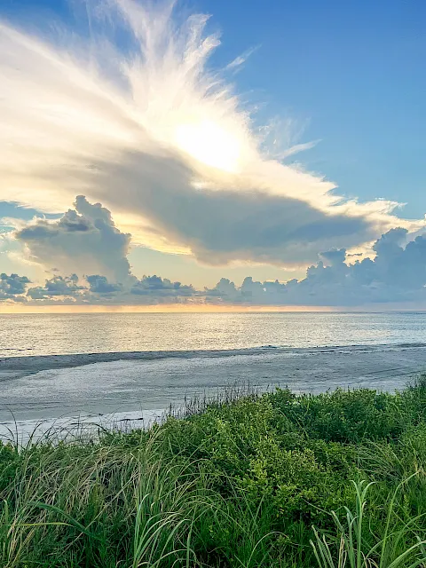 Sunrise over a tranquil ocean beach, with green grasses in the foreground and golden rays cutting through dramatic clouds.