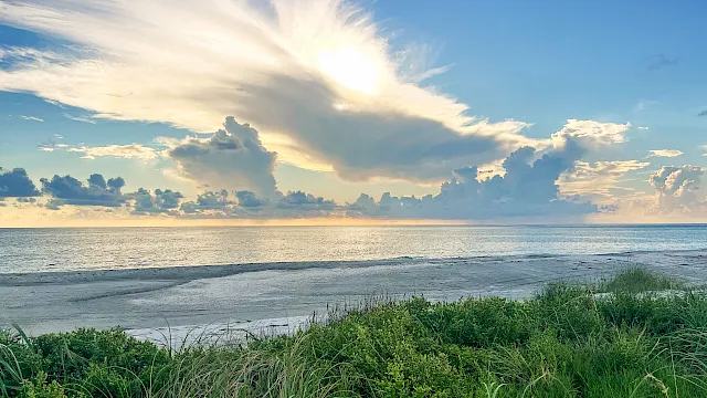 Sunrise over a tranquil ocean beach, with green grasses in the foreground and golden rays cutting through dramatic clouds.