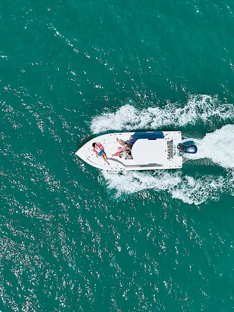 An aerial view of a white speedboat cutting across teal ocean water, tossing up a foamy wake as people enjoy the ride.