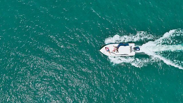 An aerial view of a white speedboat cutting across teal ocean water, tossing up a foamy wake as people enjoy the ride.
