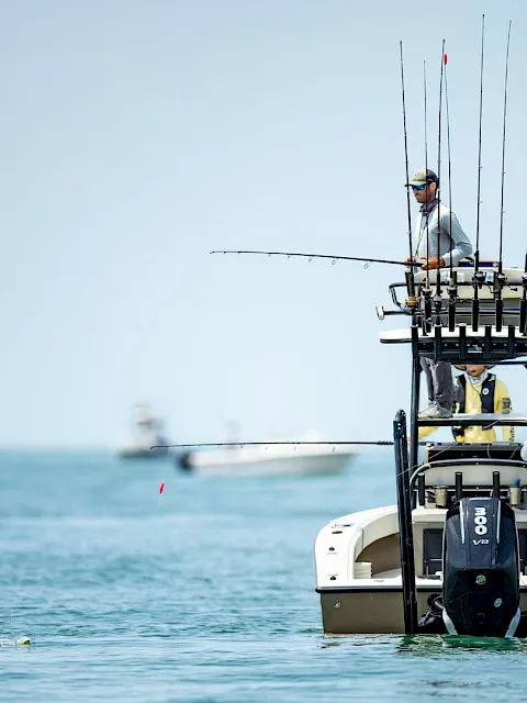 A fisherman in sunglasses stands on a small boat, steering while multiple fishing rods rise above the sea.