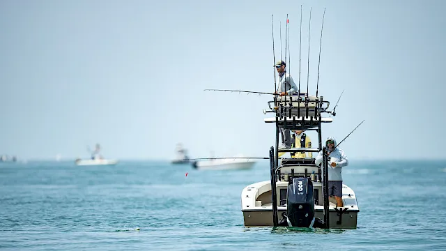 A fisherman in sunglasses stands on a small boat, steering while multiple fishing rods rise above the sea.