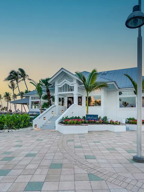 White resort building with steps, palm trees, and a sunset sky along a paved walkway beside a lamp post.
