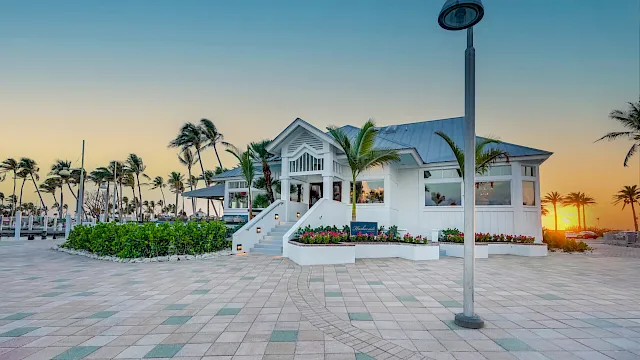 White resort building with steps, palm trees, and a sunset sky along a paved walkway beside a lamp post.