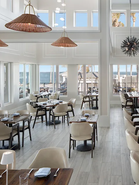 Bright seaside restaurant dining room with set tables, cushioned chairs, hanging rattan lamps, and ocean-view windows.