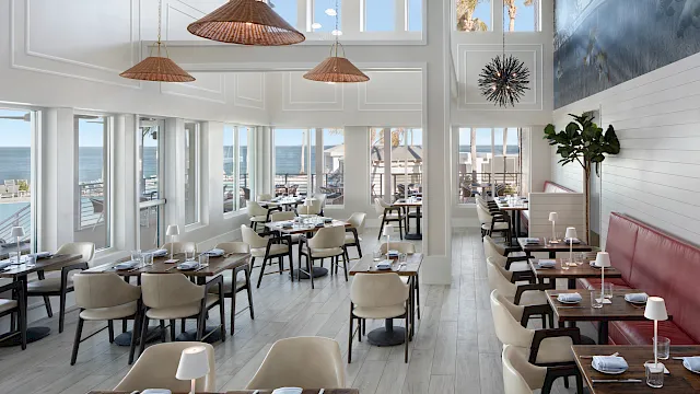 Bright seaside restaurant dining room with set tables, cushioned chairs, hanging rattan lamps, and ocean-view windows.