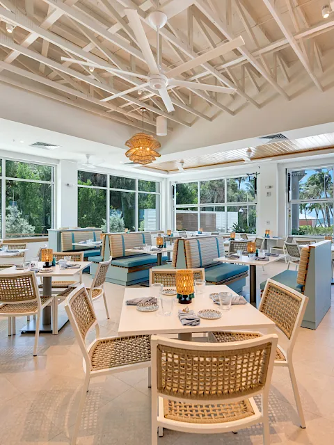 A bright waterfront dining area with neatly set tables, rattan pendant lights, and large windows showing palm trees and the sea.