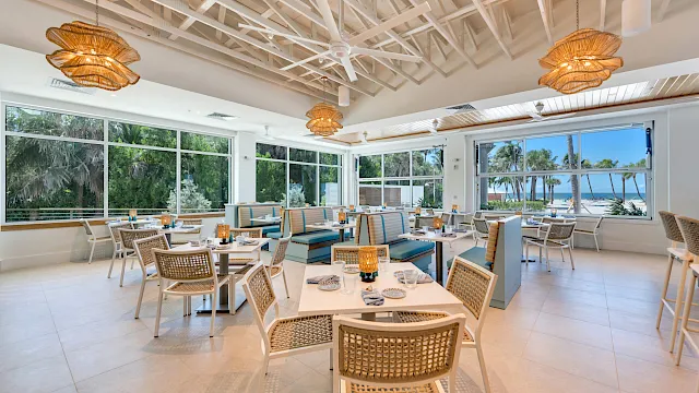 A bright waterfront dining area with neatly set tables, rattan pendant lights, and large windows showing palm trees and the sea.