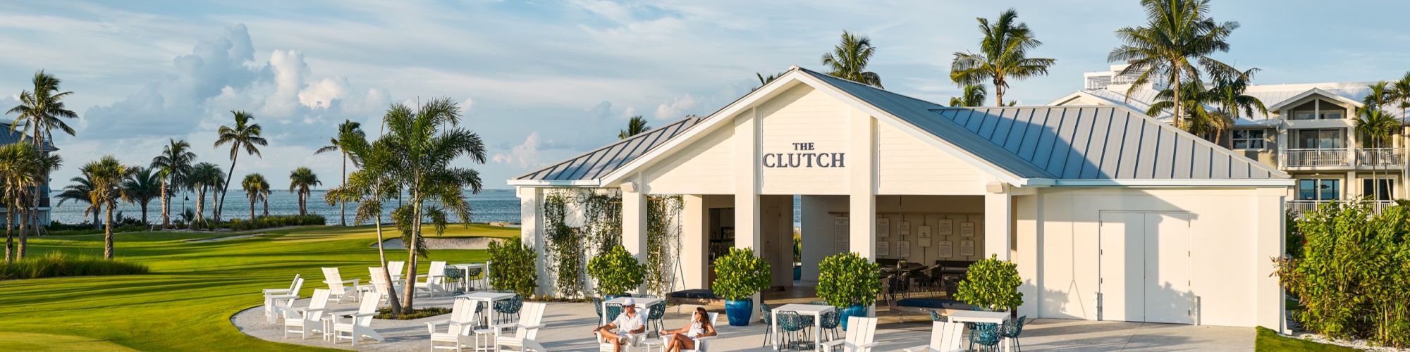 A tropical seaside resort lounge called &ldquo;THE CLUTCH&rdquo; with palm trees, white chairs, green lawns, and a blue-sky view.