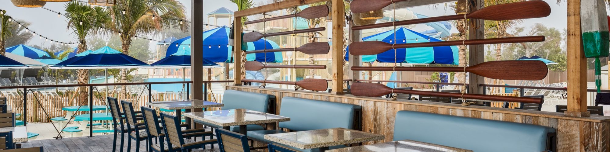 Resort bar patio with wood-and-bamboo tiki d&eacute;cor, hanging lights, cushioned booths, and caf&eacute; tables under blue umbrellas.