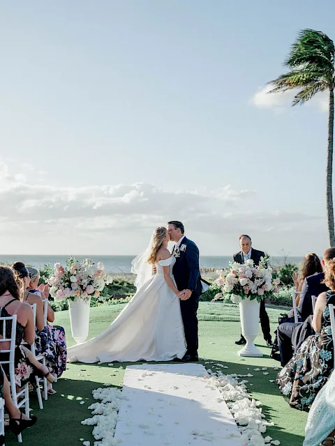 A beach wedding ceremony with a bride and groom kissing at the altar, surrounded by guests and a tall palm under sunny skies.