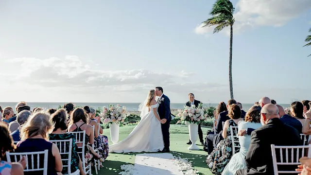 A beach wedding ceremony with a bride and groom kissing at the altar, surrounded by guests and a tall palm under sunny skies.