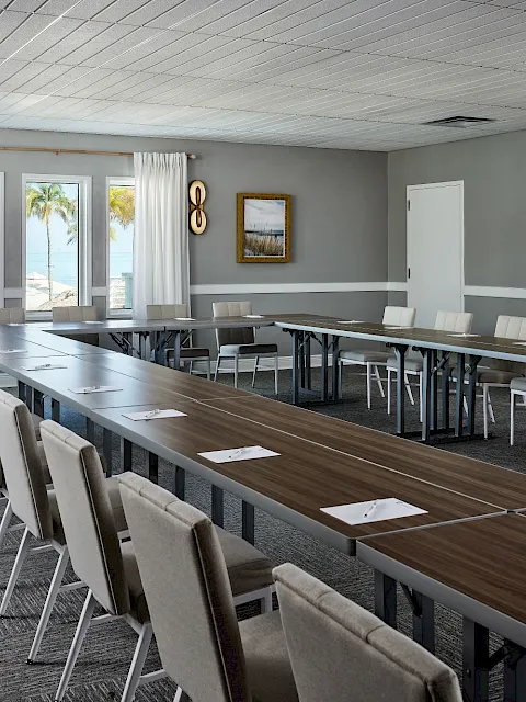 A modern conference room with a long wood table, cushioned chairs, curtains, wall art, and a bright beach-view window.