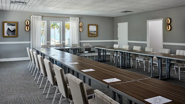 A modern conference room with a long wood table, cushioned chairs, curtains, wall art, and a bright beach-view window.