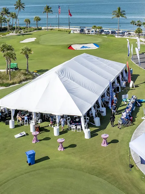 Palm-lined seaside golf course with event tents, chairs, and flags on the green beside the ocean.