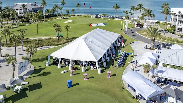 Palm-lined seaside golf course with event tents, chairs, and flags on the green beside the ocean.
