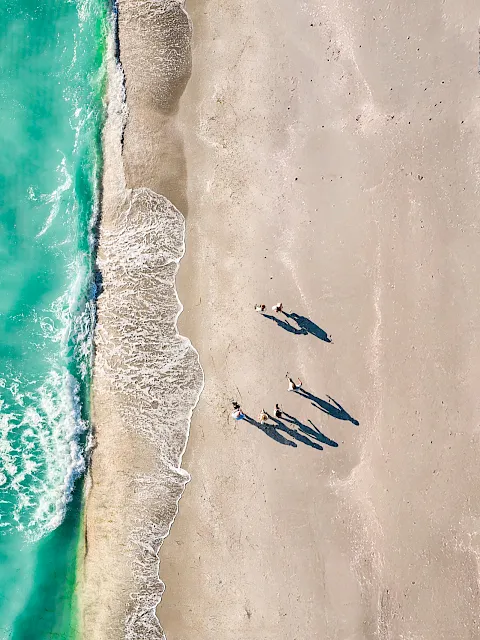 Aerial view of a sandy beach with turquoise waves, sunbathers, and rows of blue umbrellas and deck chairs.