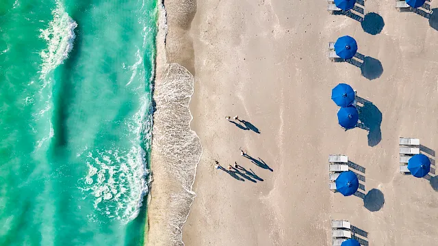 Aerial view of a sandy beach with turquoise waves, sunbathers, and rows of blue umbrellas and deck chairs.