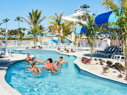 Sunny resort pool with palm trees and a big blue water slide, as guests float in inner tubes and splash in the water.