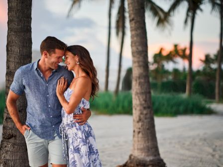 A couple share a romantic moment, hugging and smiling by palm trees on a sunny beach.