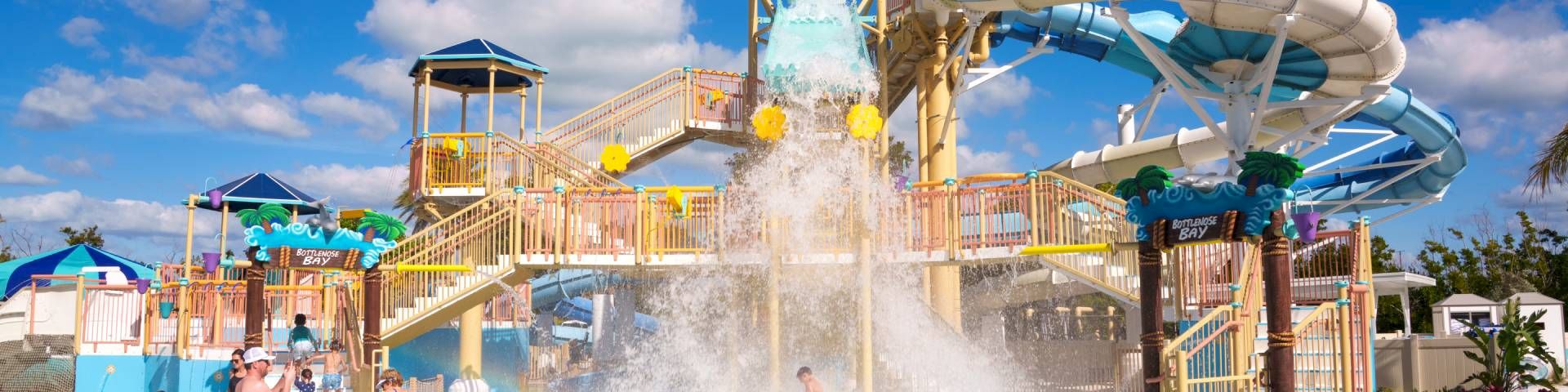 A sunny water park scene: kids and adults splash in a pool as a big slide jets water into the air.
