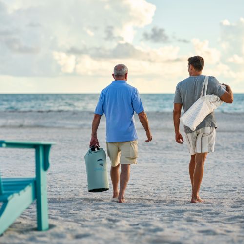 Two men walk barefoot along a sandy beach toward the ocean, carrying beach bags near a blue boardwalk chair.