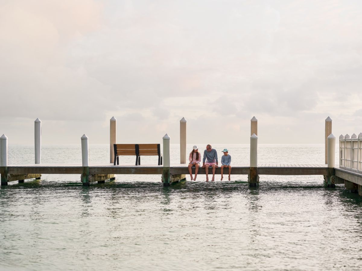 Three people relax on a seaside dock, sitting on a bench while the ocean and cloudy sky stretch behind them.