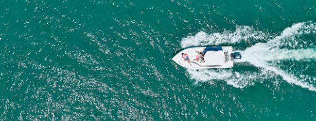 An overhead view shows a small motorboat speeding across teal water, leaving a foamy wake behind it.