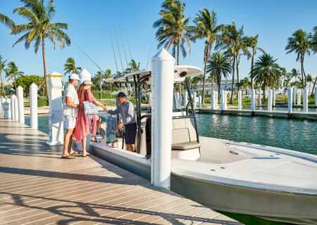 People are boarding a small motorboat at a sunny dock, surrounded by tropical palm trees and calm water.