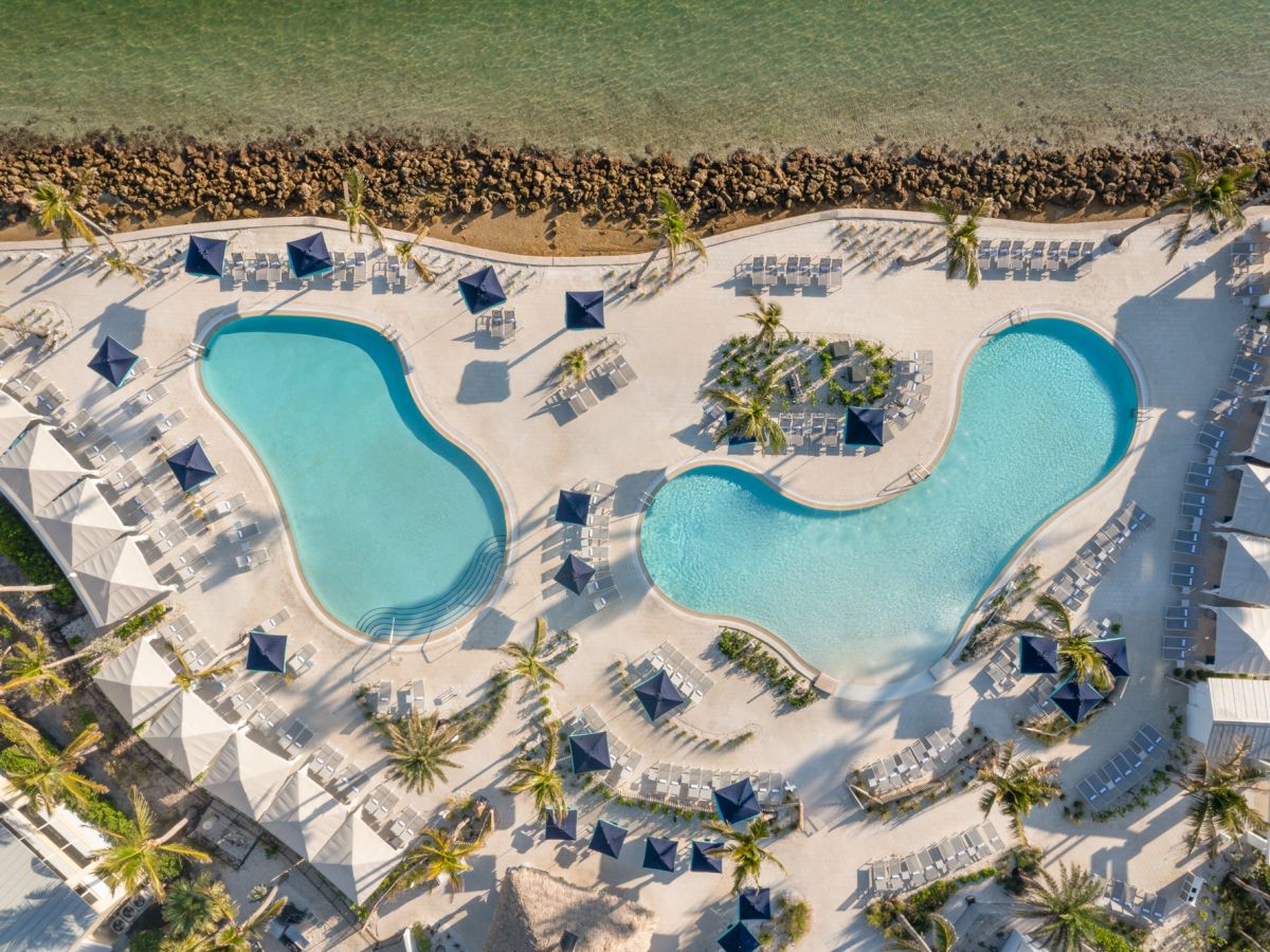 Aerial view of a tropical resort with two turquoise swimming pools, lounge chairs, umbrellas, and palm trees by the beach.