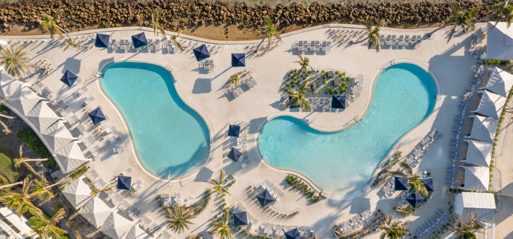 Aerial view of a tropical resort with two turquoise swimming pools, lounge chairs, umbrellas, and palm trees by the beach.