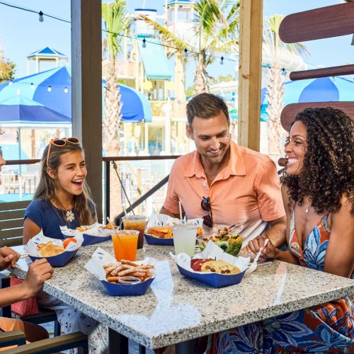 A group of friends laugh and share a sunny outdoor meal at a wooden table with drinks and burgers, celebrating together.