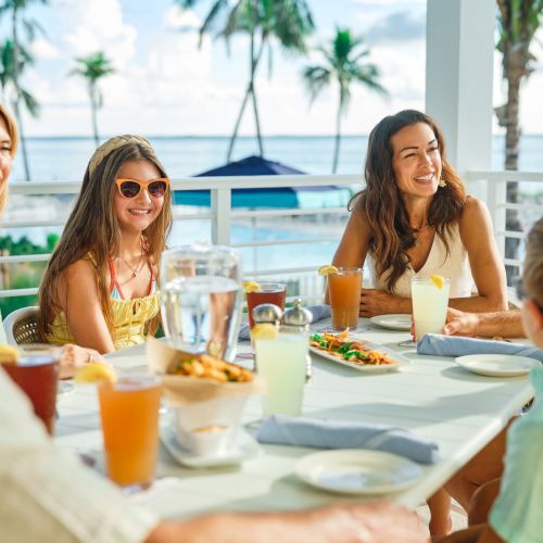 Friends dine on a sunny seaside patio, smiling and toasting with drinks in front of the ocean and palm trees.