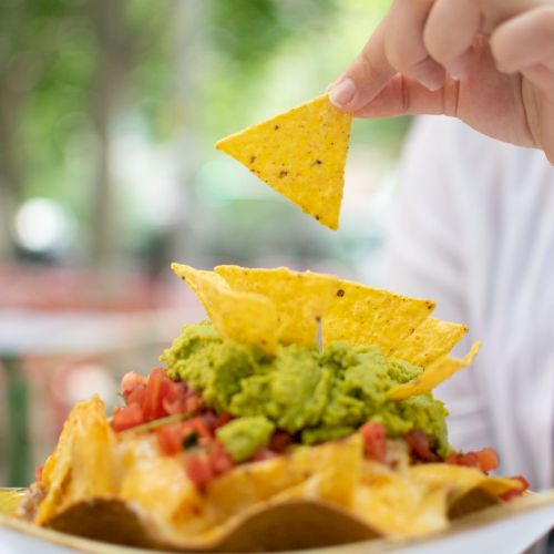 A person holds a crunchy tortilla chip above a taco bowl with guacamole, salsa, and veggies in the sunshine.