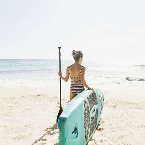 A woman sits on a beach holding a paddle beside her turquoise surfboard, facing the ocean under a clear sky.