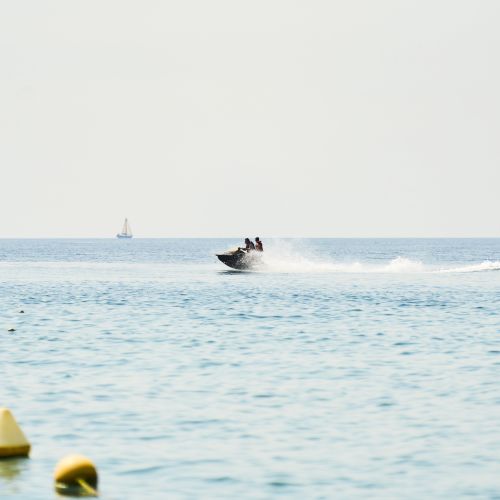 A speedboat with two riders sprays across the sea, with a distant sailboat and yellow buoys floating in the water.